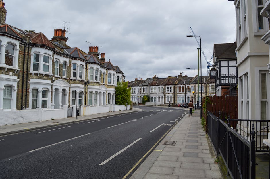 A residential street scene along Kennington Road in Kennington, featuring a row of Victorian-style terraced houses with white-painted facades, bay windows, and red-tiled roofs. The pavement is clean and well-maintained, with a black metal fence bordering some of the properties. The road is marked with white dashed lines and appears freshly cleaned, free of litter and debris. Overhead, the sky is cloudy, casting diffuse light over the quiet, tidy street. The scene includes a few pedestrians walking along the sidewalk. In the background, construction cranes are visible, indicating ongoing development in the area. The overall impression emphasizes cleanliness, urban residential character, and maintained streets, aligning with the services offered by Cleaners Kennington for surface cleaning and maintenance of flats on Kennington Road.