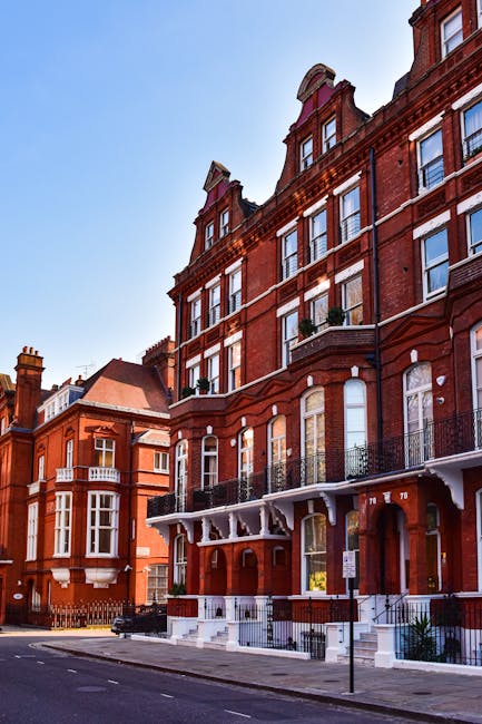 A row of red-brick residential buildings on Kennington Road viewed from the street during daytime. The buildings feature large sash windows, some with small balconies with black wrought iron railings, and ornamental architectural details. The pavement in front is clean, with a streetlamp and a parking sign visible. The sky is clear and blue, highlighting the well-maintained exterior of the buildings. Cleaners Kennington provides comprehensive cleaning services to maintain the appearance and hygiene of such residential properties, including surface cleaning and deep sanitation of common areas.