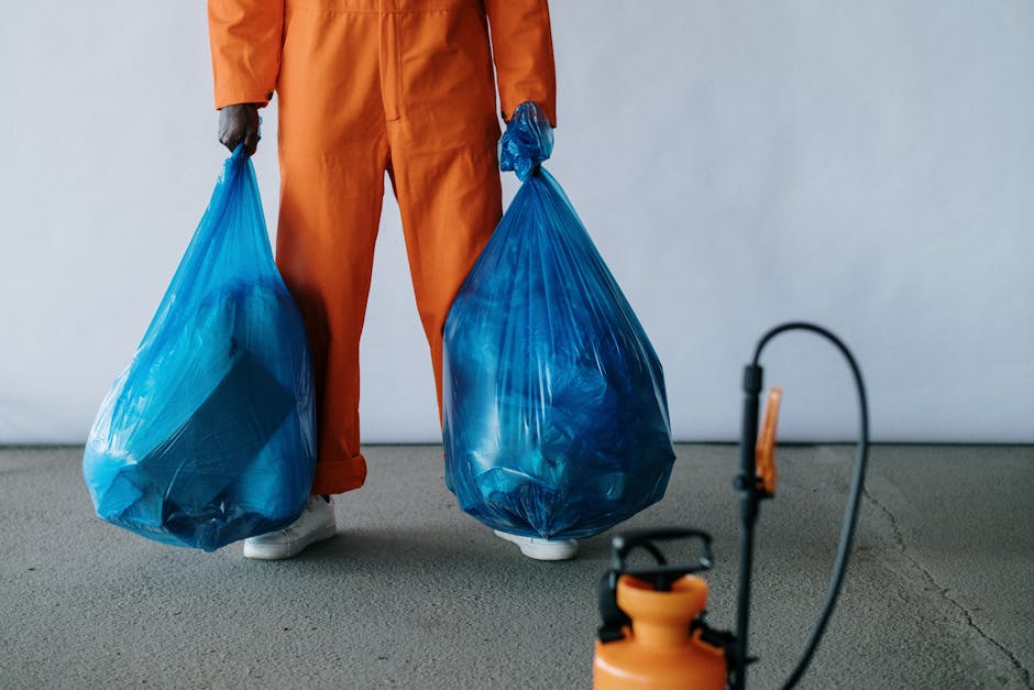 A street-level view of a municipal street cleaner from Cleaners Kennington dressed in an orange high-visibility uniform with reflective stripes, standing on a paved sidewalk near a building entrance. 
