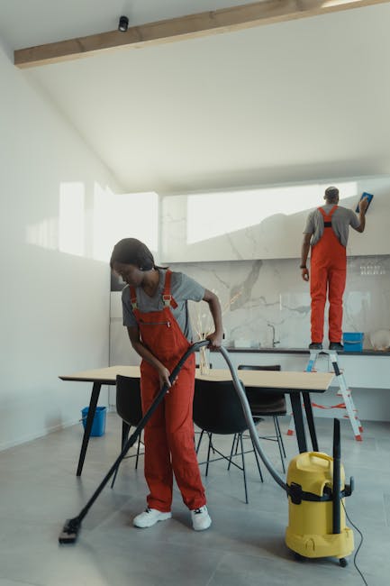 Two professional cleaners from Cleaners Kennington are performing surface cleaning in a modern kitchen. The cleaner in the foreground is using a vacuum cleaner on the tiled floor, which appears freshl